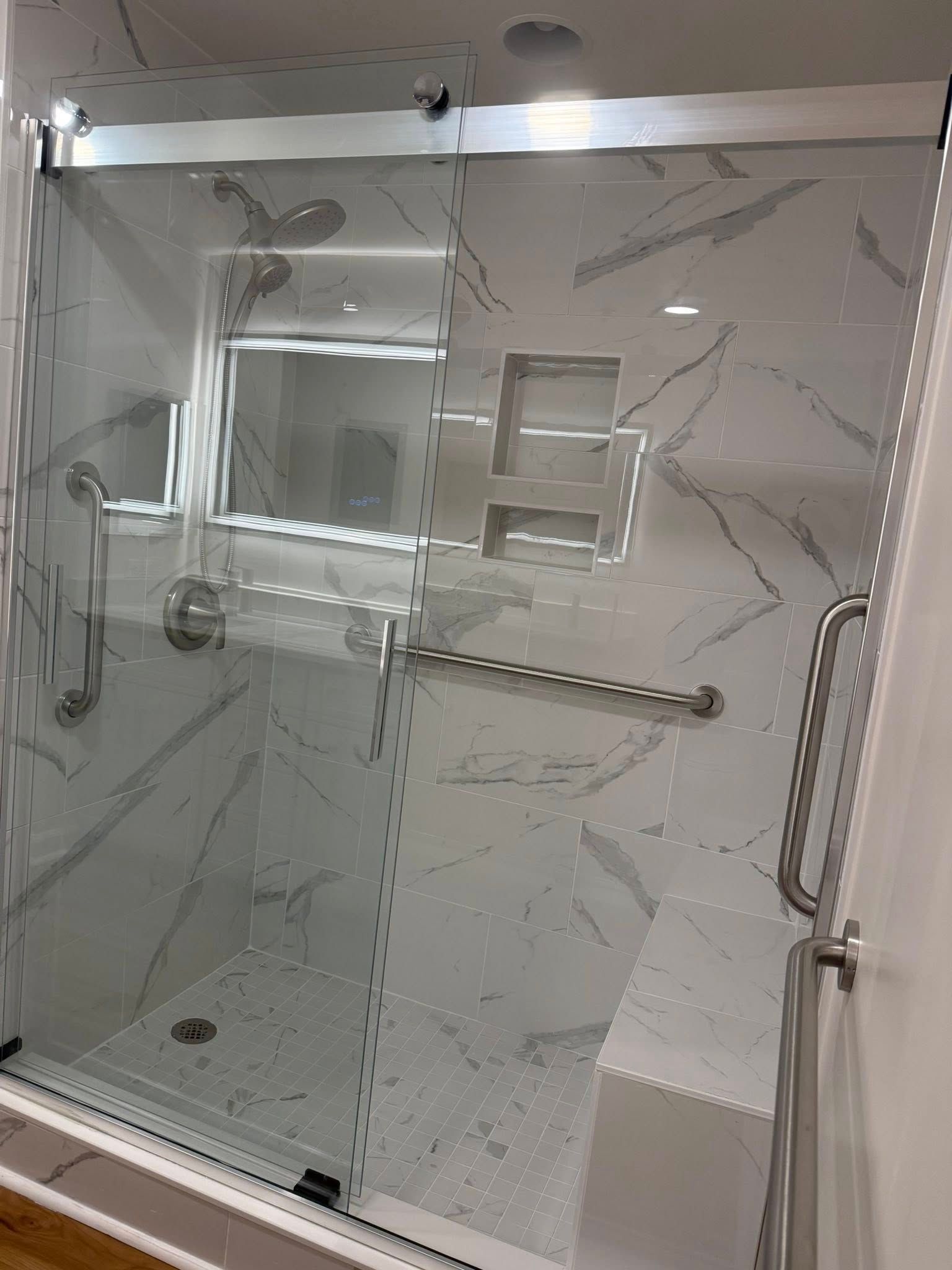 A walk-in shower with marble-patterned tiles, a built-in seat, multiple safety grab bars, and a sliding glass door.