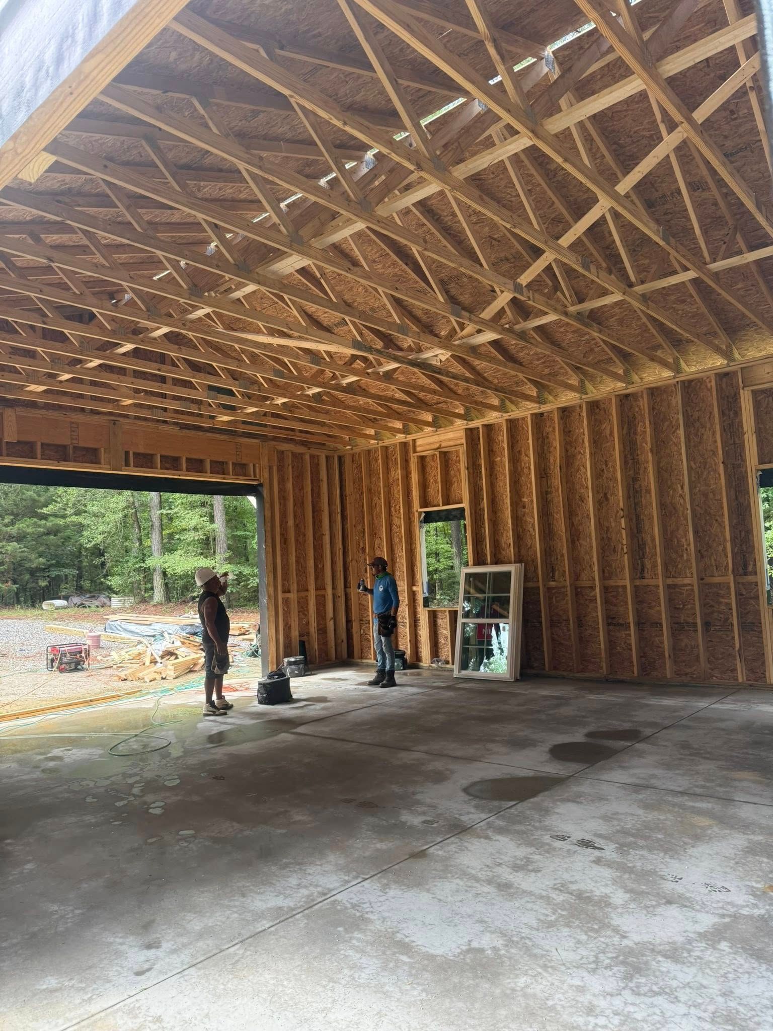 Two people stand inside the unfinished wooden frame of a garage under construction with an open doorway facing woods.