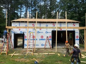 Construction workers build a single-story structure with white house wrap, surrounded by ladders and wooden framing poles.