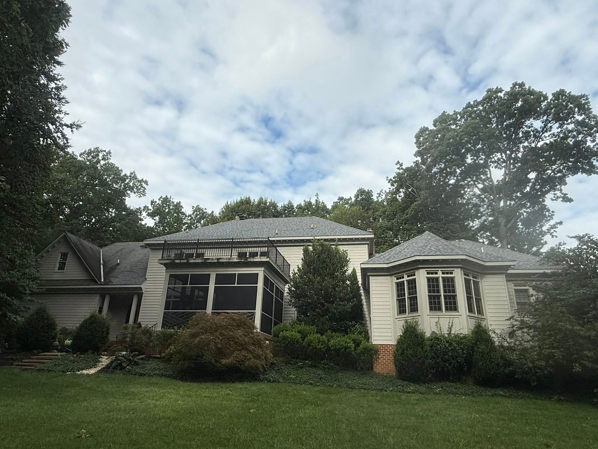 A beige, multi-gabled house with a sunroom, surrounded by lush trees under a cloudy sky.