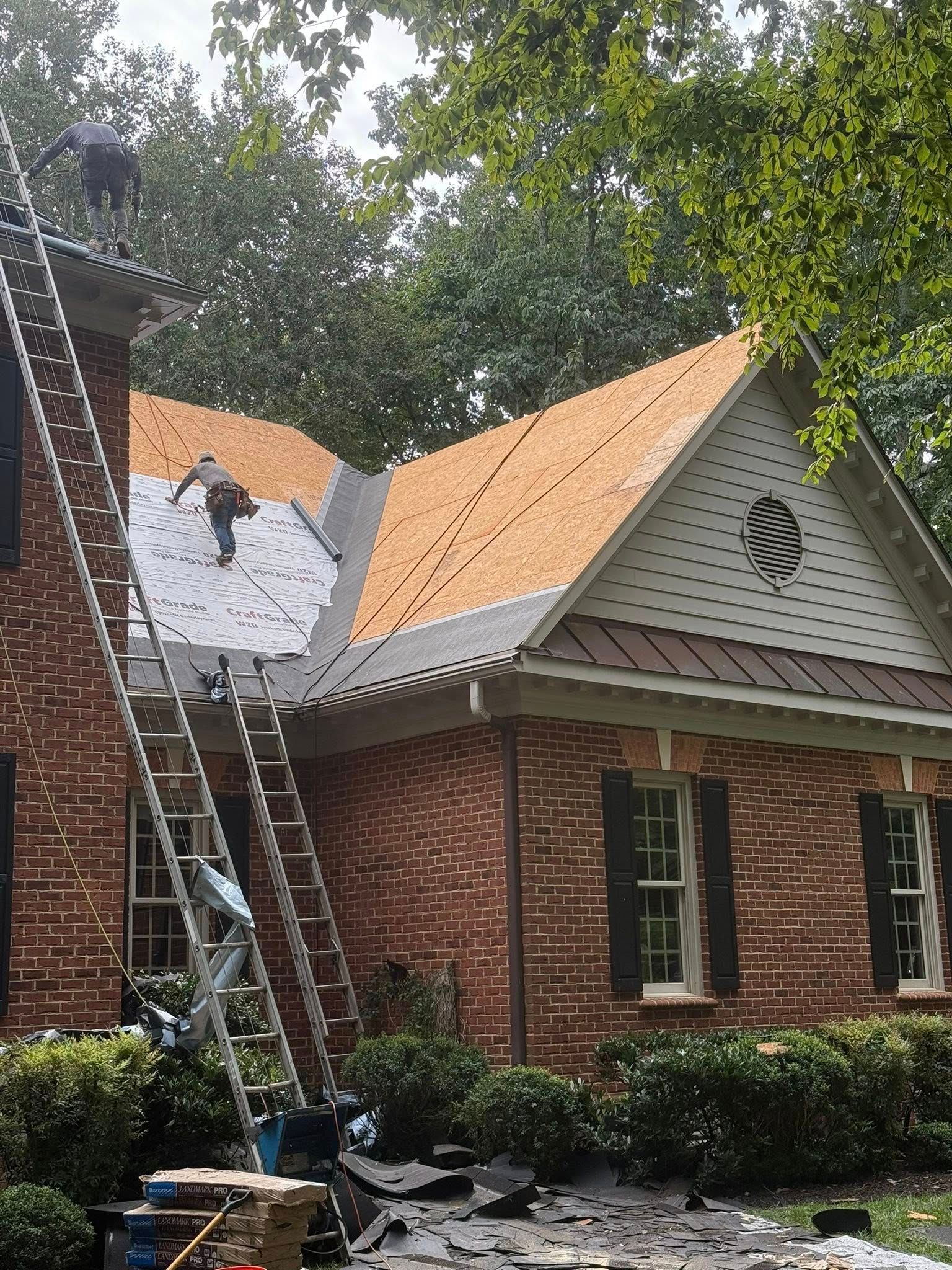 Workers repair the roof of a two-story brick home, applying underlayment over exposed wooden roof decking.