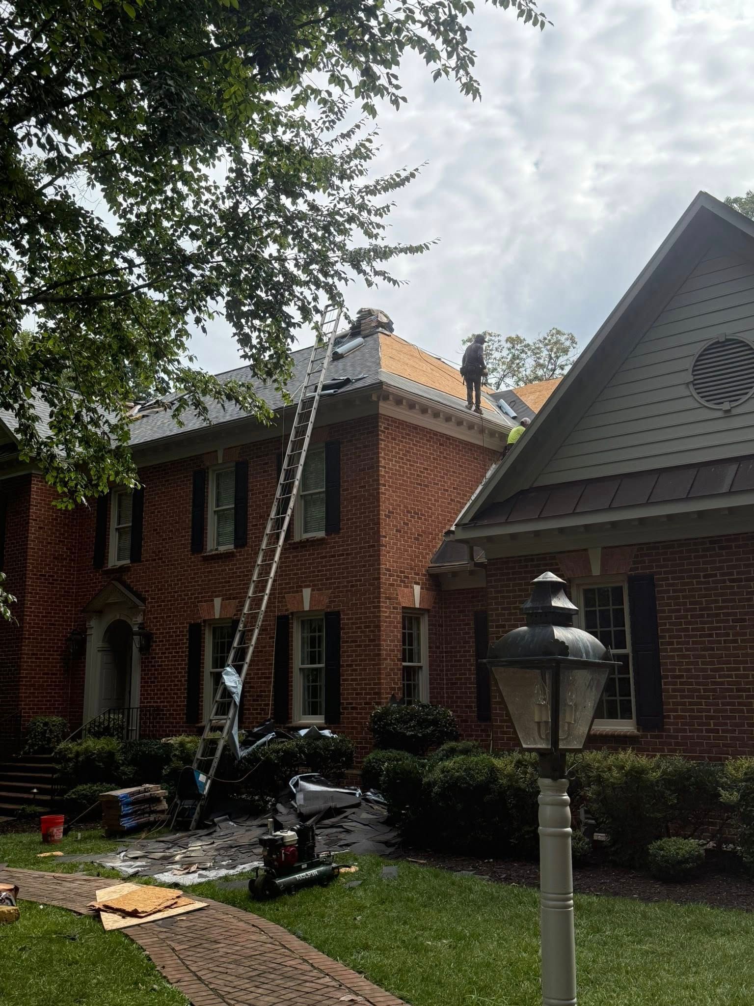 Workers on a roof, accessible by a tall ladder, repairing shingles on a two-story brick home.