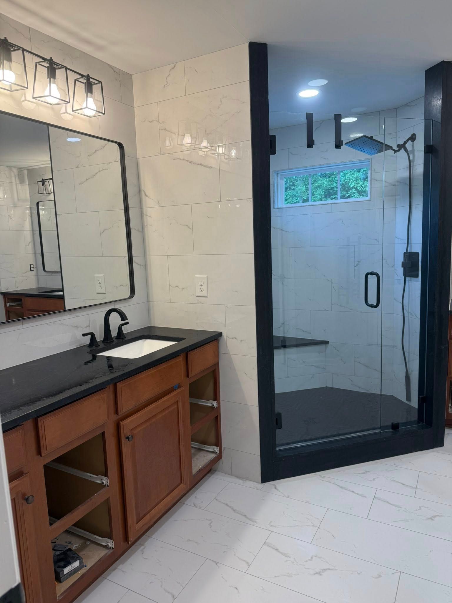 A modern bathroom with a wooden vanity, a black countertop and mirror, and a glass-enclosed shower with white tiled walls.