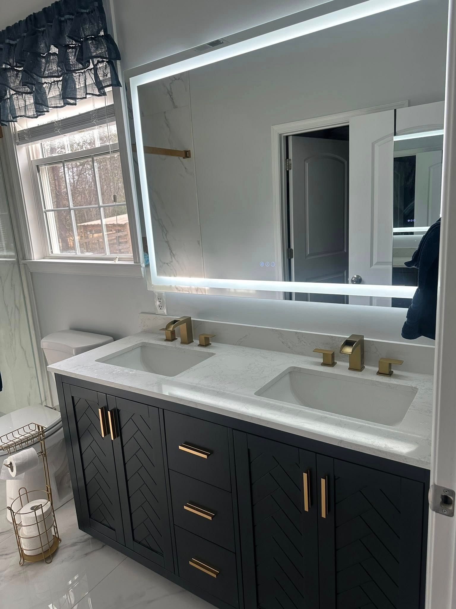 A modern bathroom featuring a black double-sink vanity with gold hardware, a white marble countertop, and a lighted mirror.