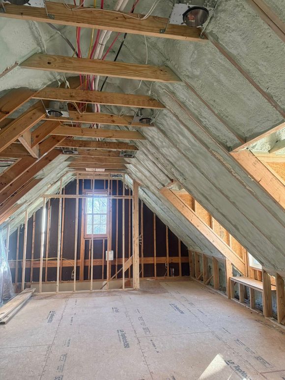 An unfinished attic with exposed wooden rafters, spray foam insulation, framing for a partition wall, and a window.