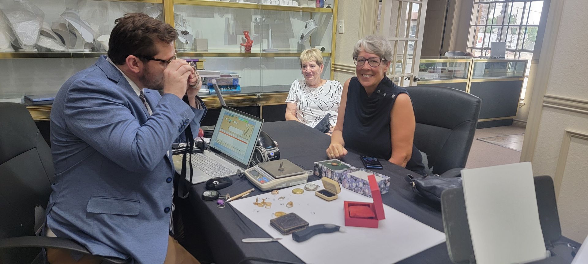 A man in a blue blazer uses a magnifying glass to inspect jewelry at a desk with two women in a jewelry store.