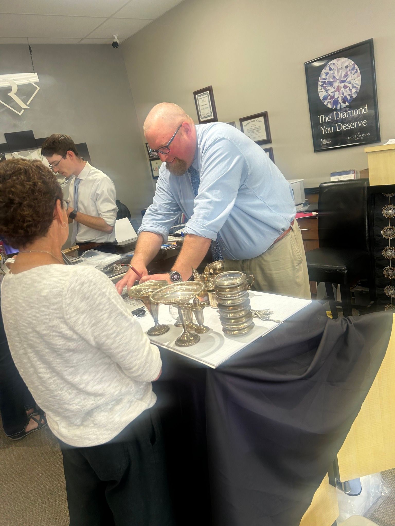 A man in a light blue shirt examines objects on a table, conversing with a woman in a white sweater. A young man in a tie stands behind them.