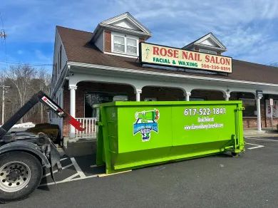 A green dumpster is parked in front of a rose nail salon.