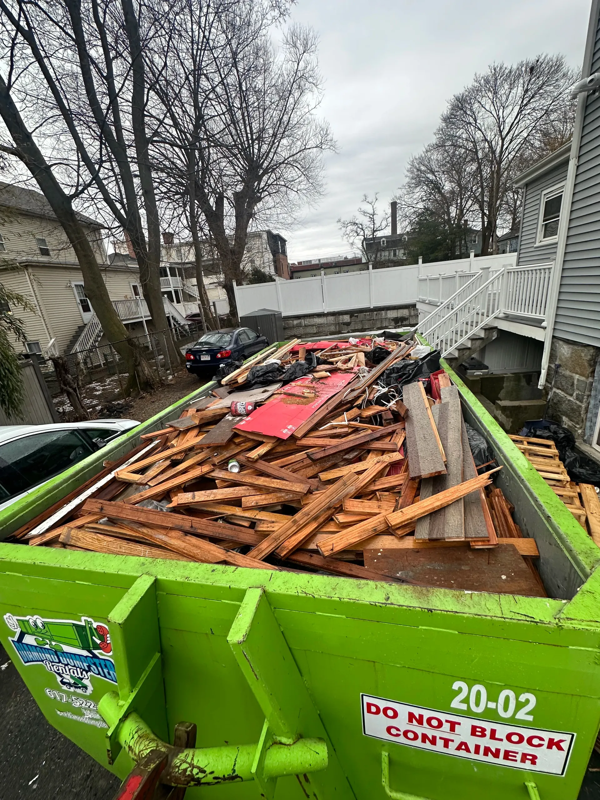 A green dumpster filled with wood is parked in front of a house.