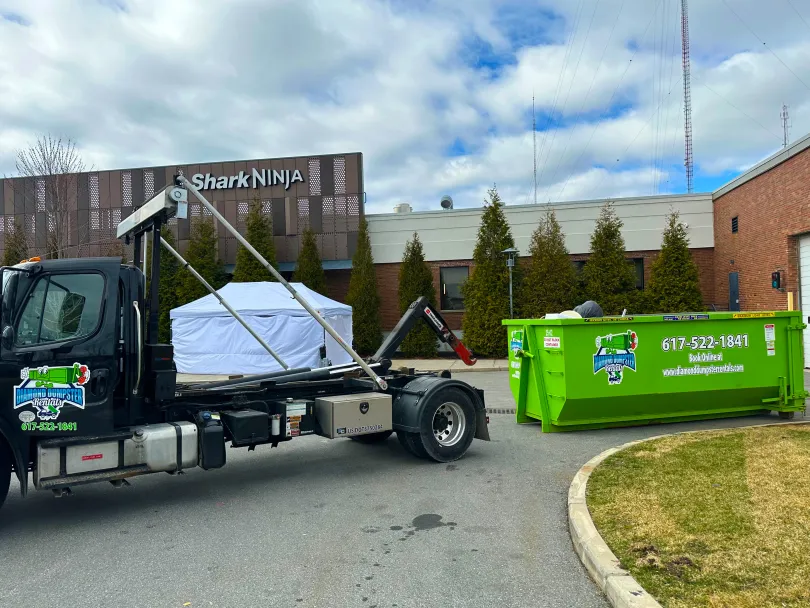 A dumpster is being towed by a truck in front of a building.