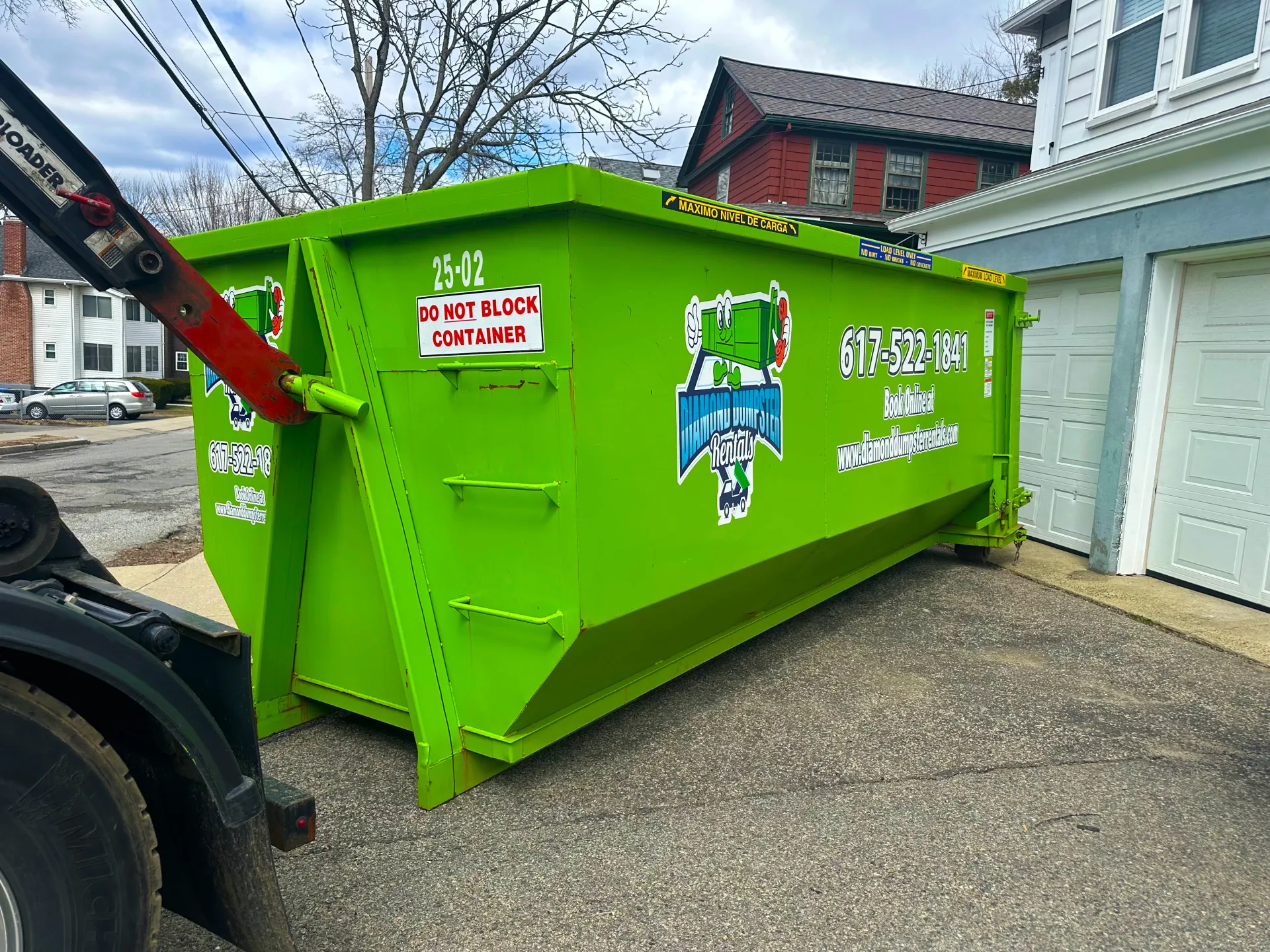 A green dumpster is sitting on the side of a truck in front of a house.