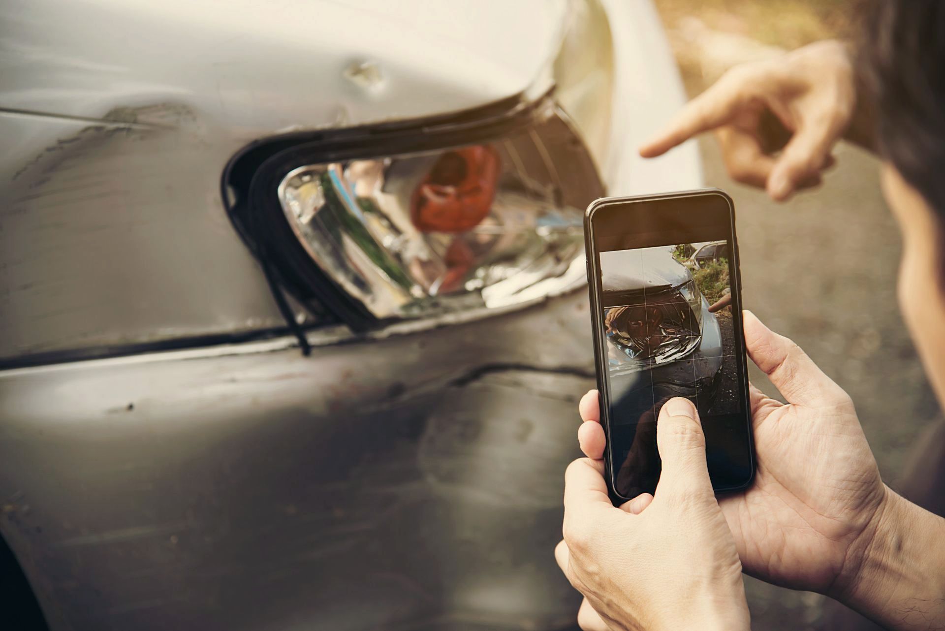 Person taking photo of car damage with a smartphone after a collision; another person points to the damage.