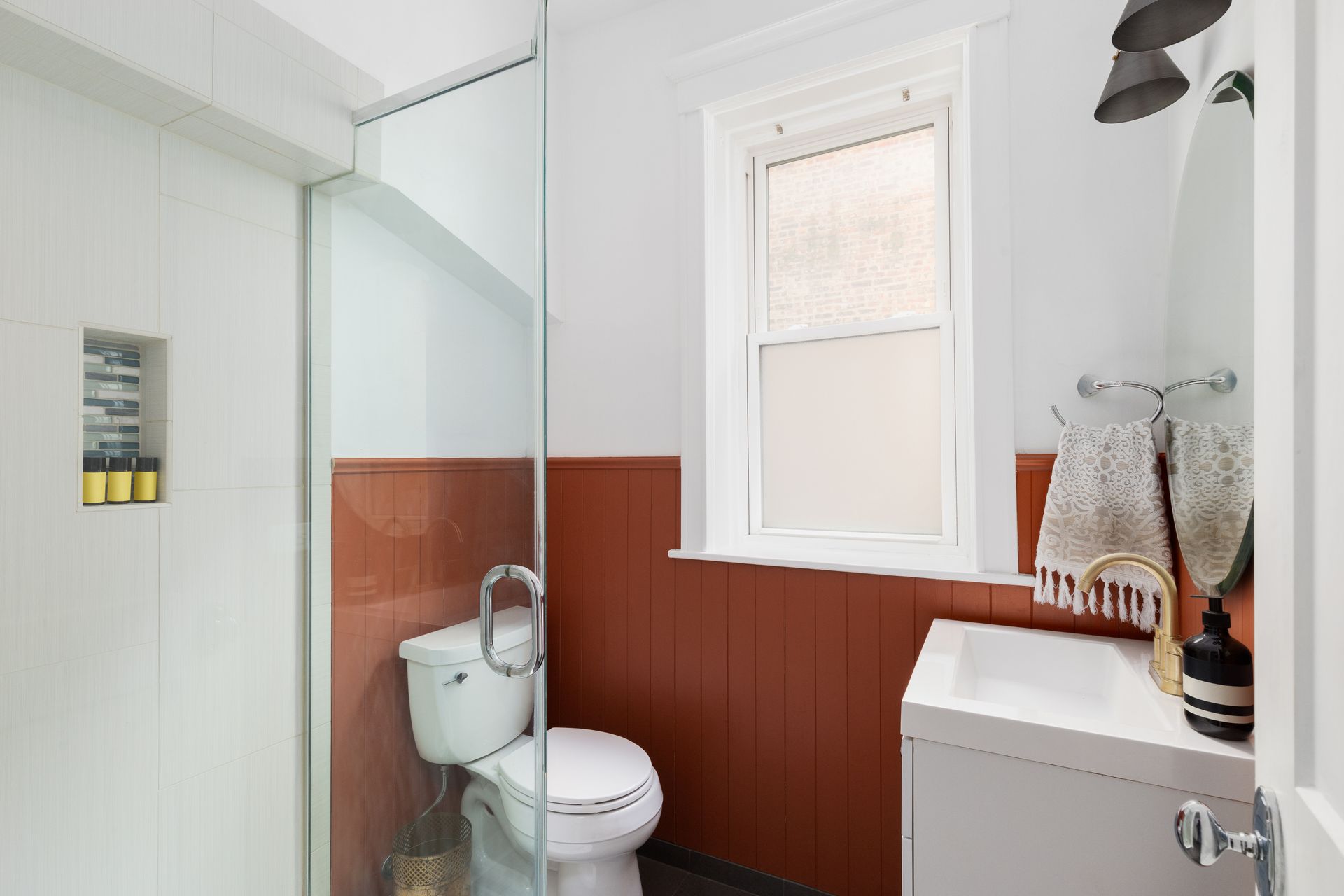A modern bathroom with a glass shower, white walls, and warm rust-colored wainscoting below a window.