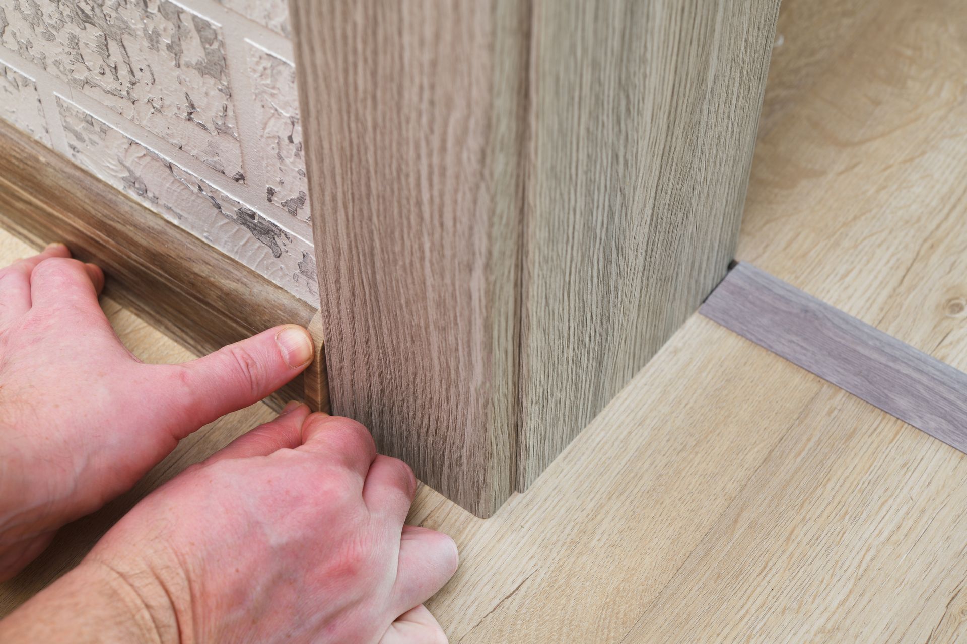 Hands fitting a wood-patterned baseboard against a vertical trim piece on a light-colored floor.