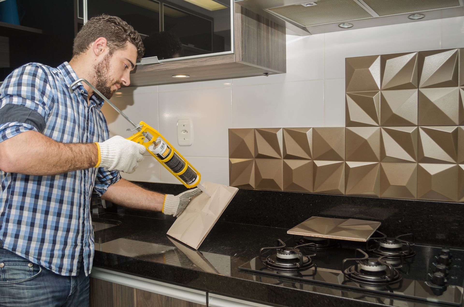 A person wearing work gloves uses a caulk gun to apply adhesive to a textured wall tile in a kitchen.