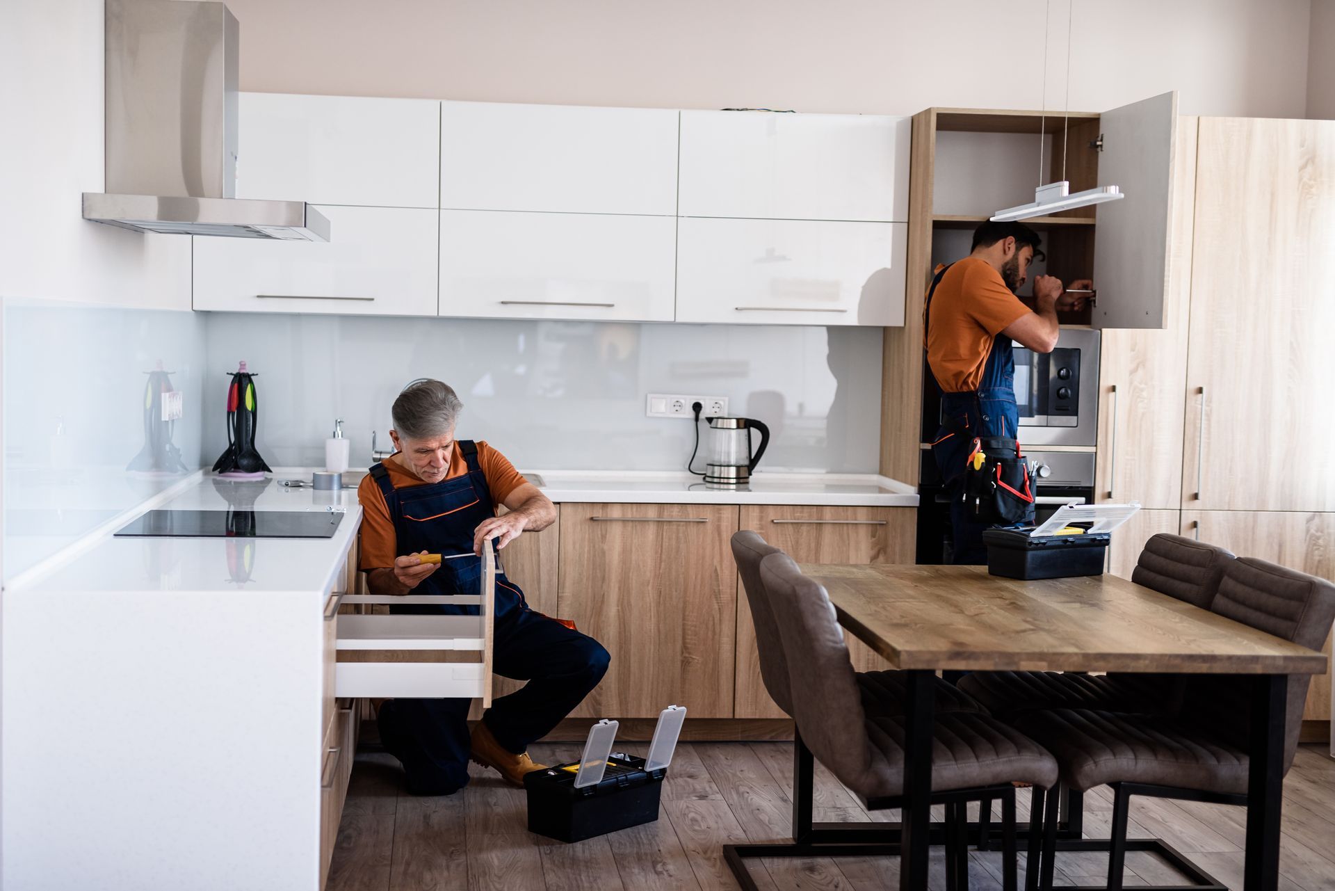 Two professionals in work clothes perform maintenance and installation tasks in a modern kitchen with white cabinetry.