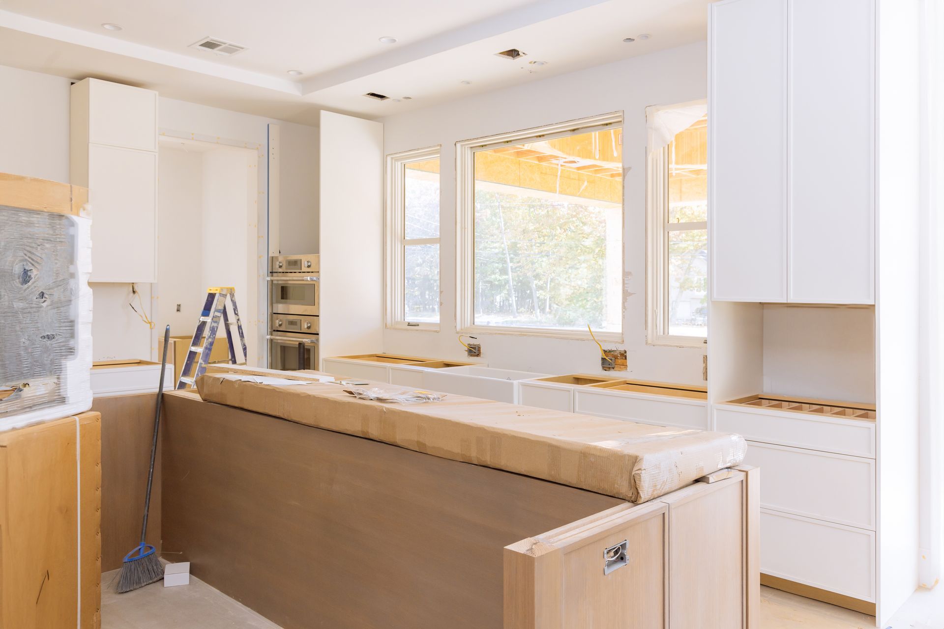 A kitchen under construction featuring light wood cabinets, white upper units, a large island, and windows facing outside.