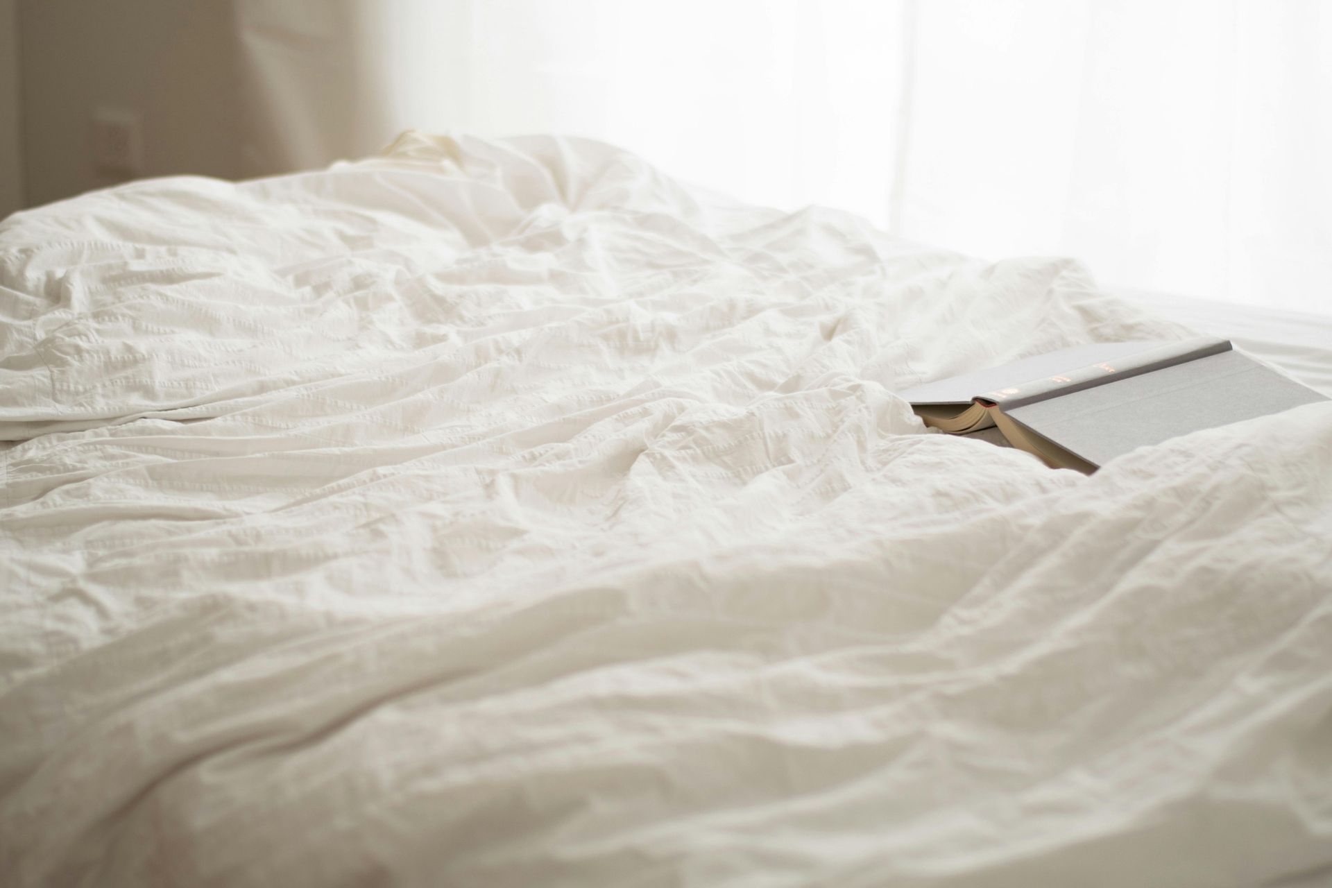 Wrinkled white bedsheets with a book lying open; soft natural light.