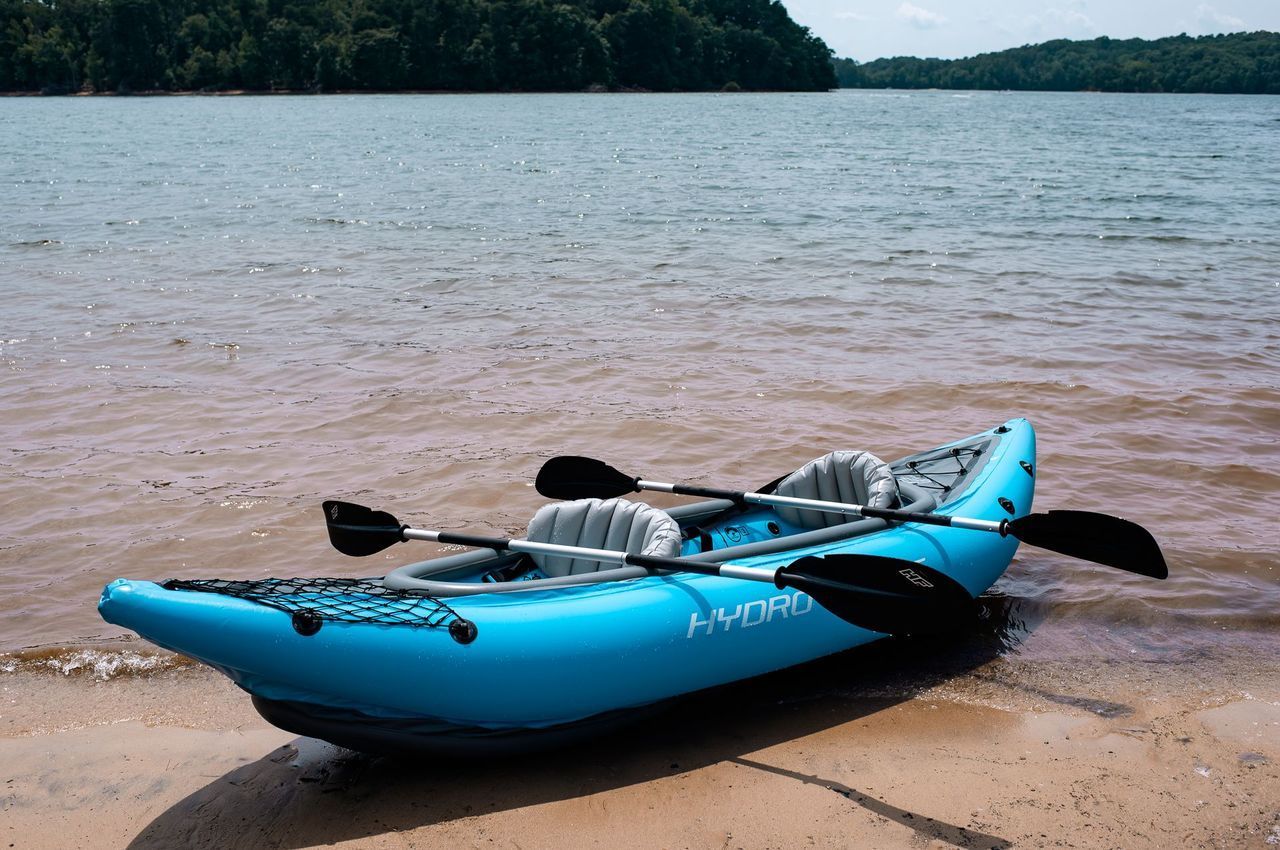 A blue kayak with two paddles is sitting on the shore of a lake.