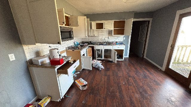 Kitchen in disrepair, cabinets open, appliances exposed, trash on floor, wood flooring.