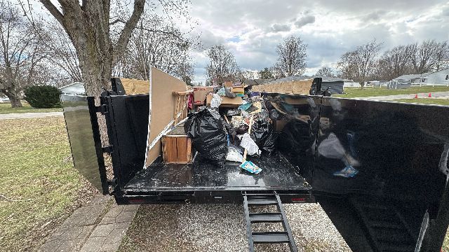 A black trailer filled with trash, including bags, cardboard, and debris, sitting outside on a lawn.