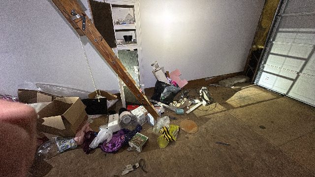Cluttered, dirty room with scattered boxes, trash, and exposed wall framing near a garage door.