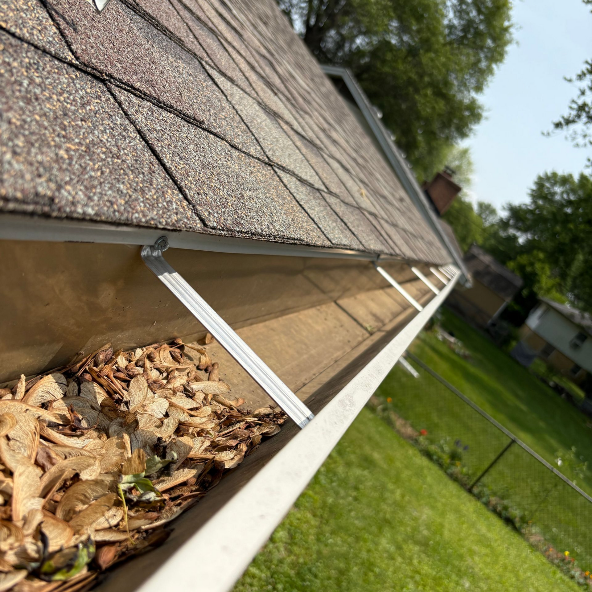 Gutter filled with leaves on a house roof, viewed from a low angle, with a green lawn in the background.