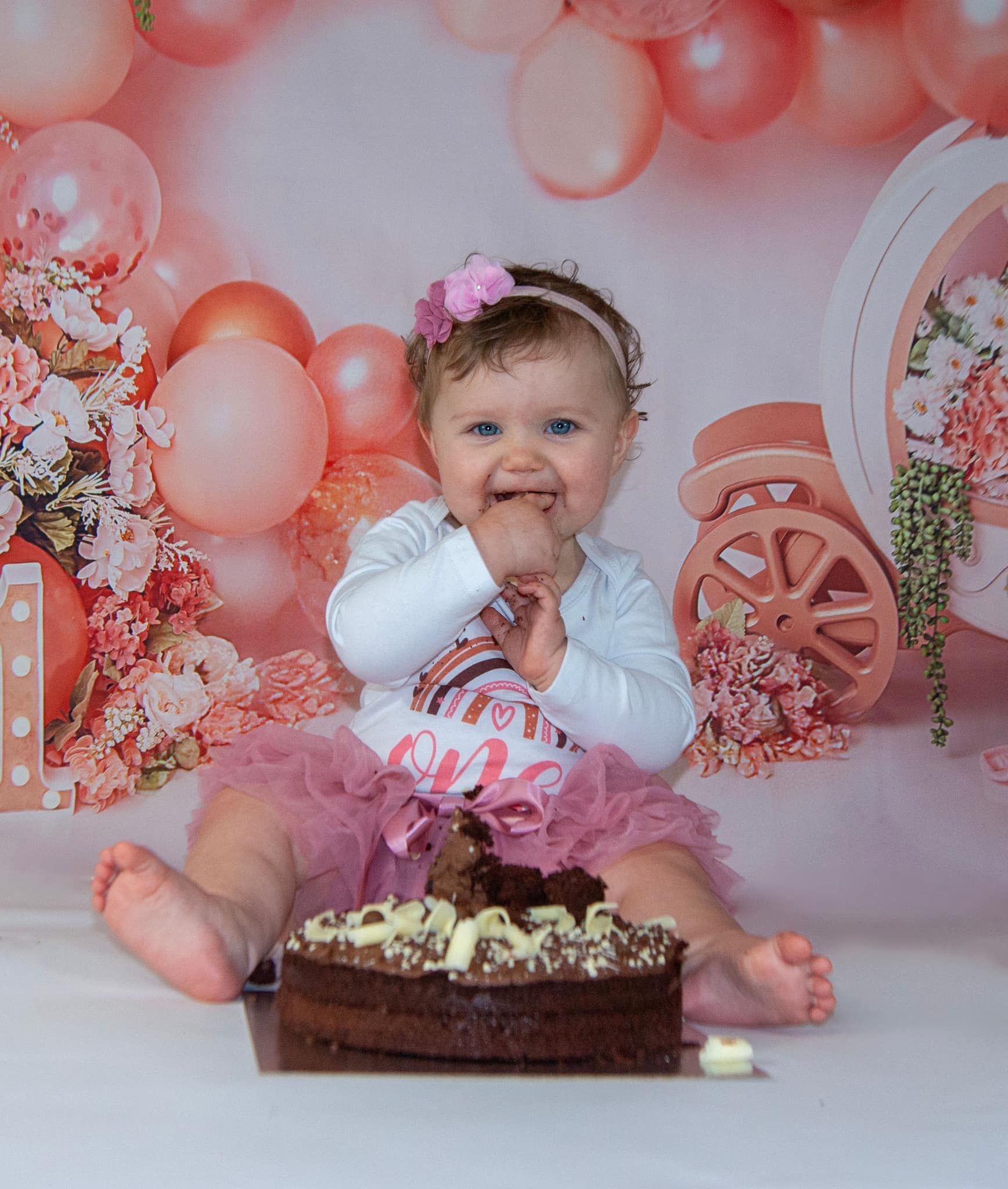 A baby girl is sitting in front of a cake with balloons in the background.