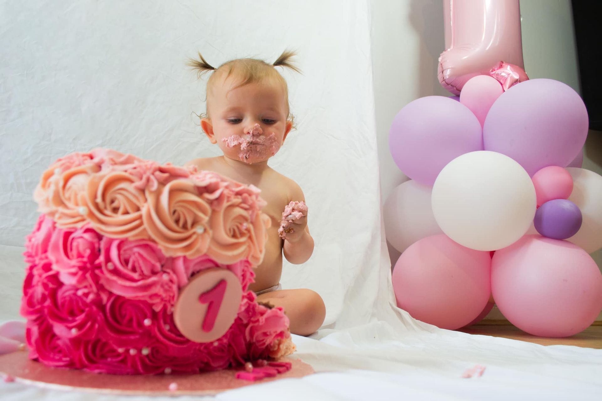 A baby is sitting next to a pink cake with balloons in the background.