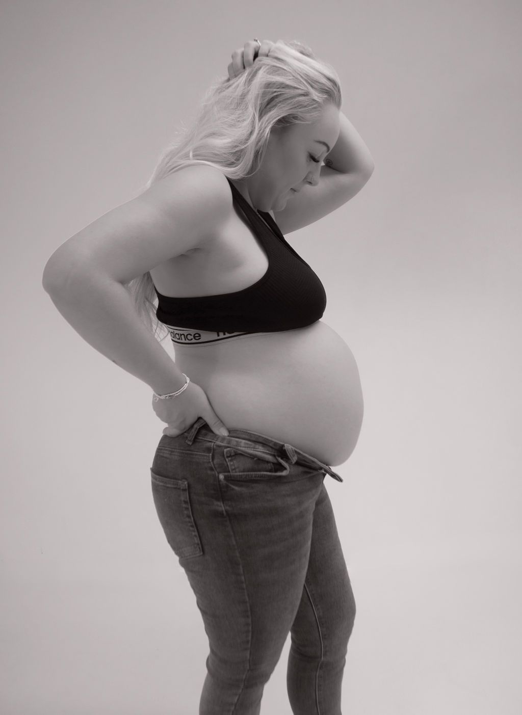 A pregnant woman in a black bra and jeans is standing with her hands on her hips.