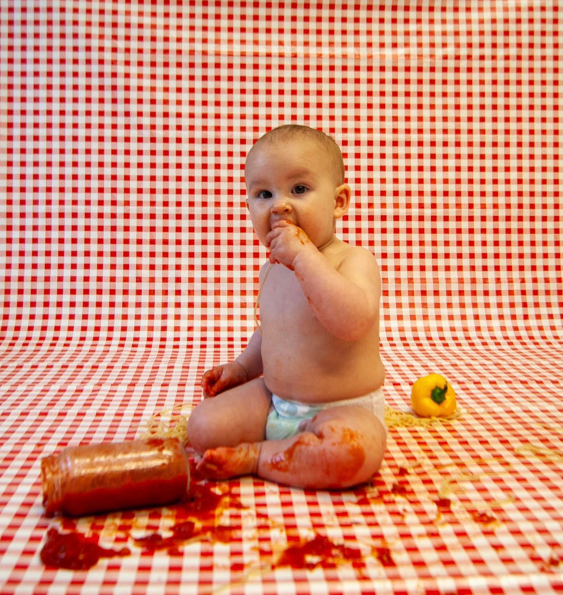 A baby in a diaper is sitting on a checkered tablecloth eating tomato sauce.