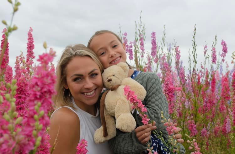 A woman and a little girl are holding a teddy bear in a field of pink flowers.