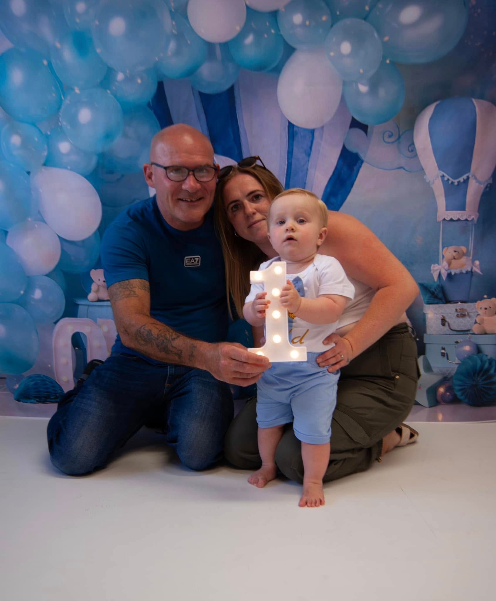 A man and woman are kneeling down next to a baby holding a lighted number one sign.