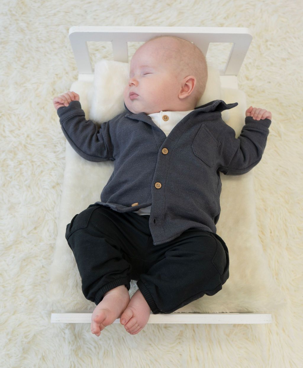 A baby wearing a grey cardigan and black pants is laying on a white bed