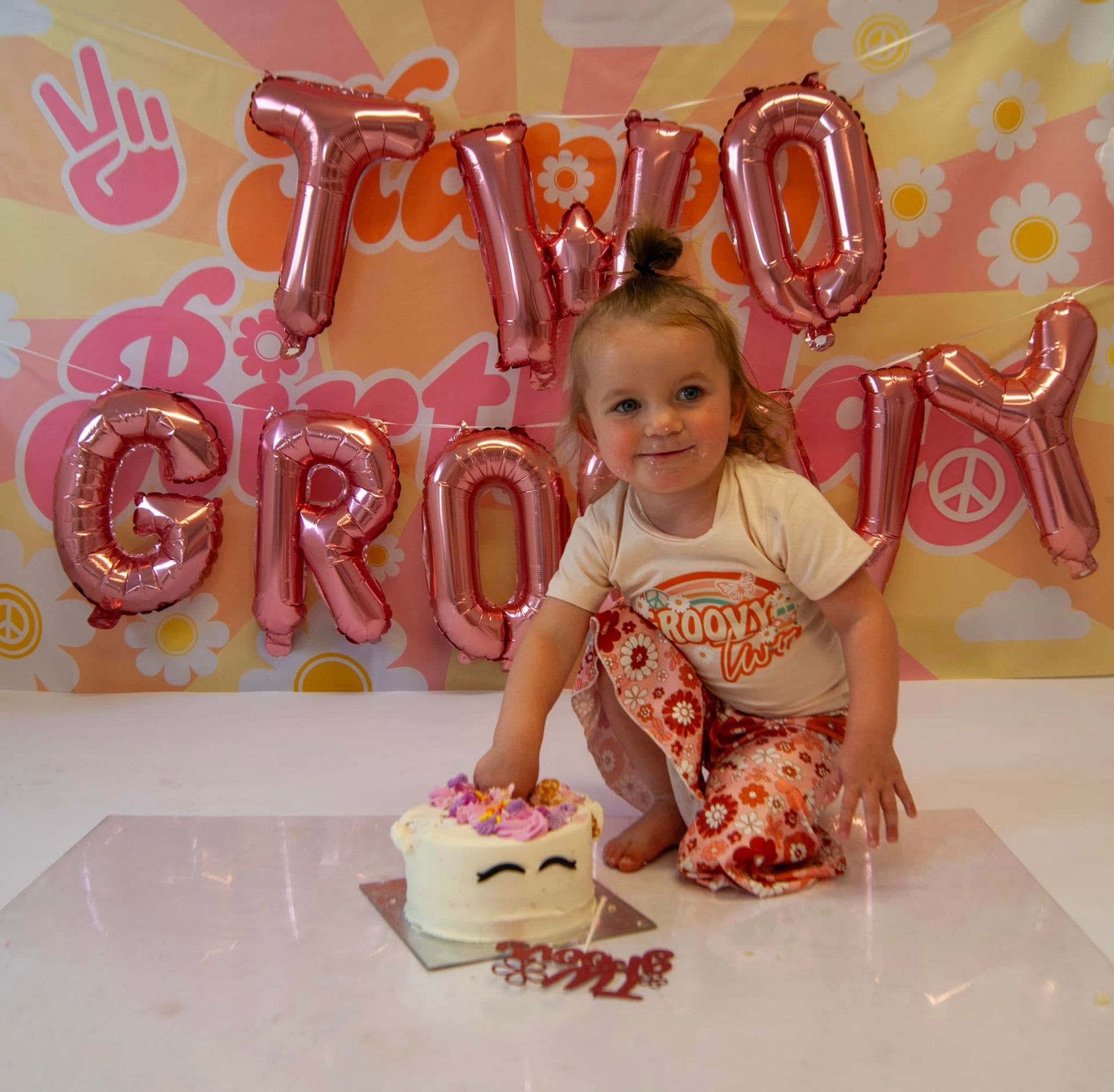 A little girl kneeling next to a cake in front of a sign that says two groovy