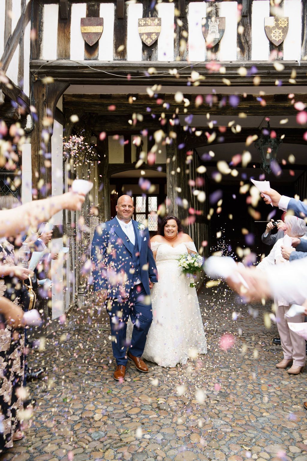 Newlyweds exit a building, showered with confetti. The couple, a man in a blue suit and a woman in a white wedding dress, smile.