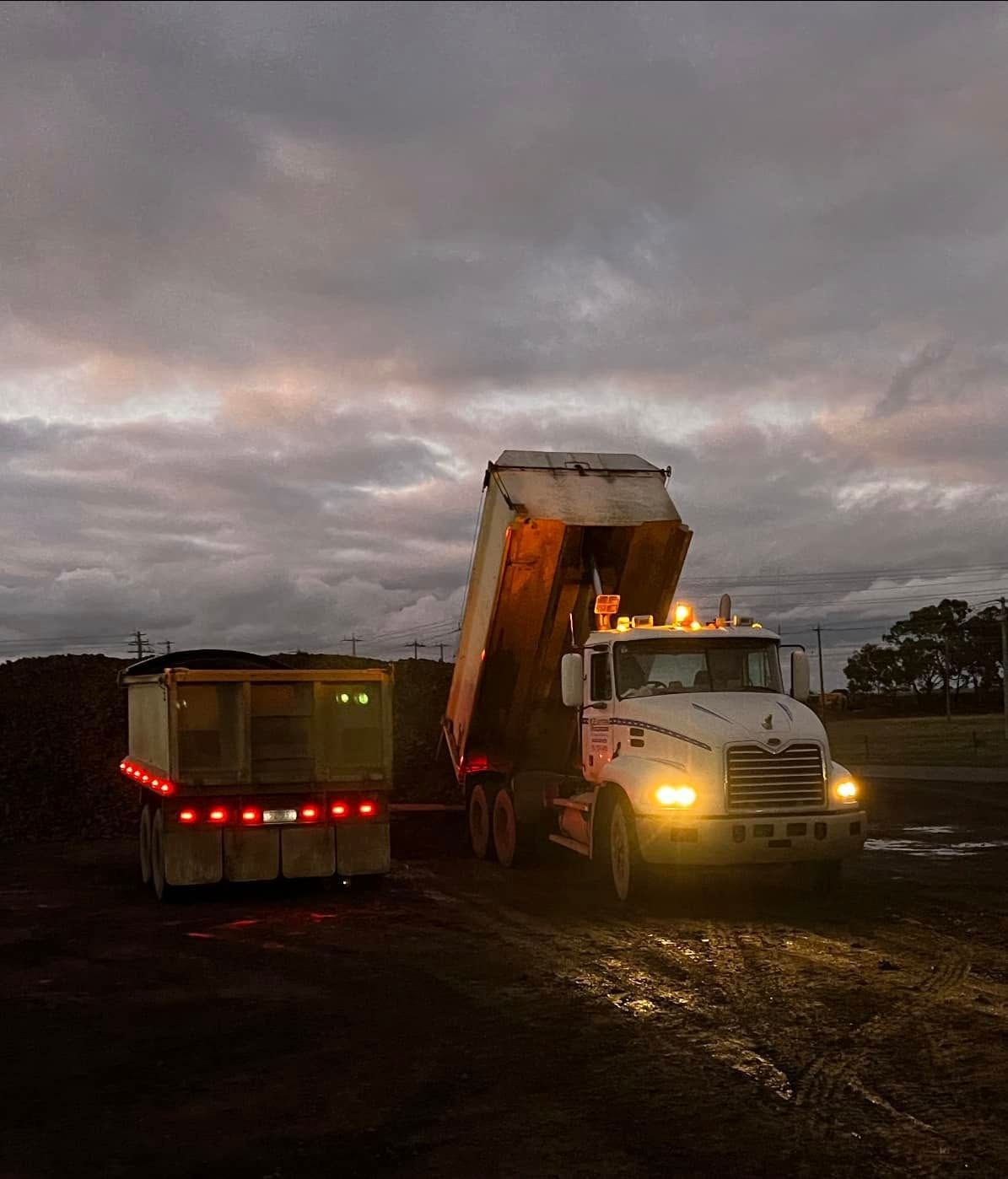 Dump truck unloading soil into a trailer at dusk on a muddy worksite.