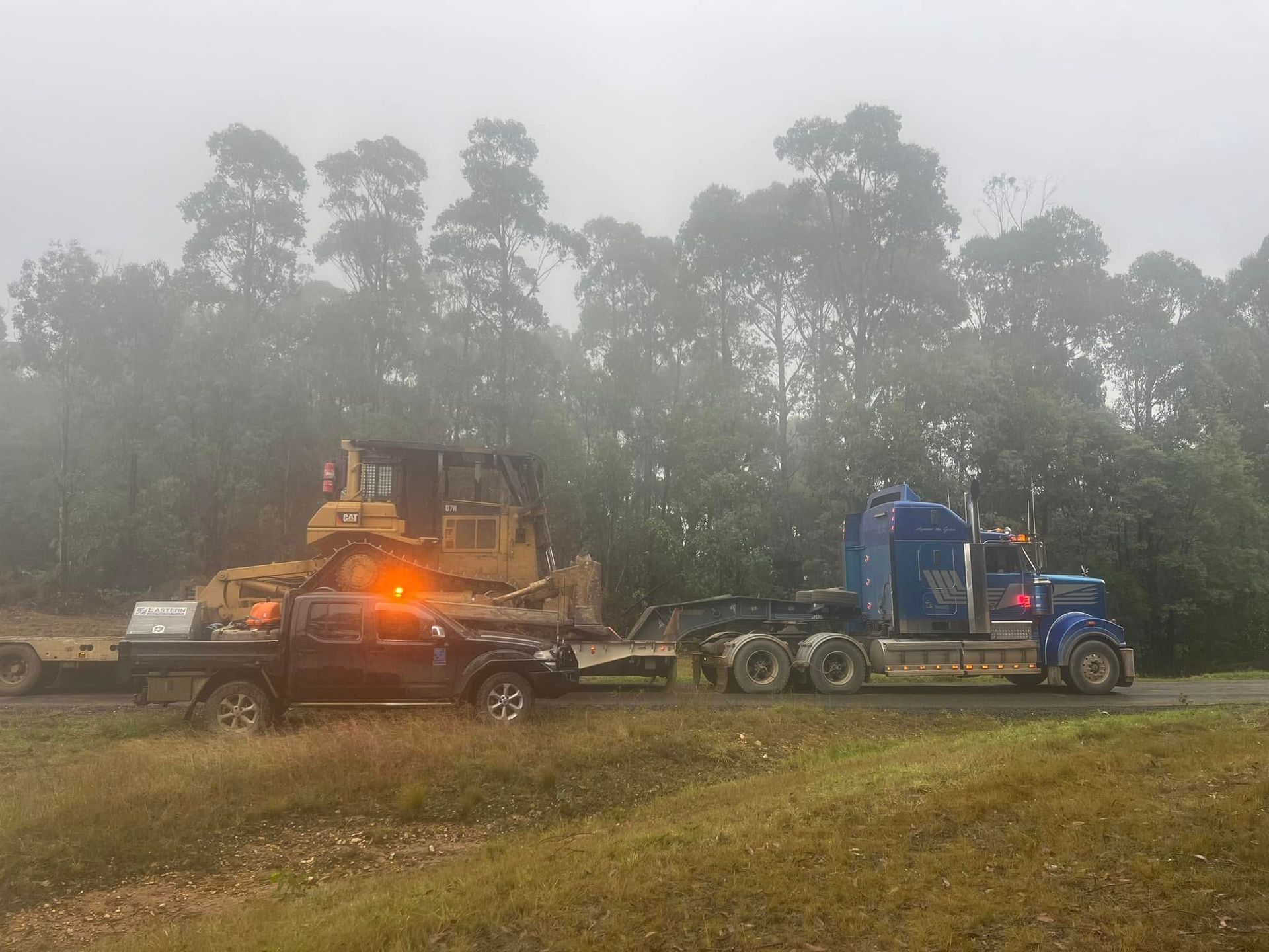 Blue truck hauling heavy machinery on a flatbed in a foggy wooded field
