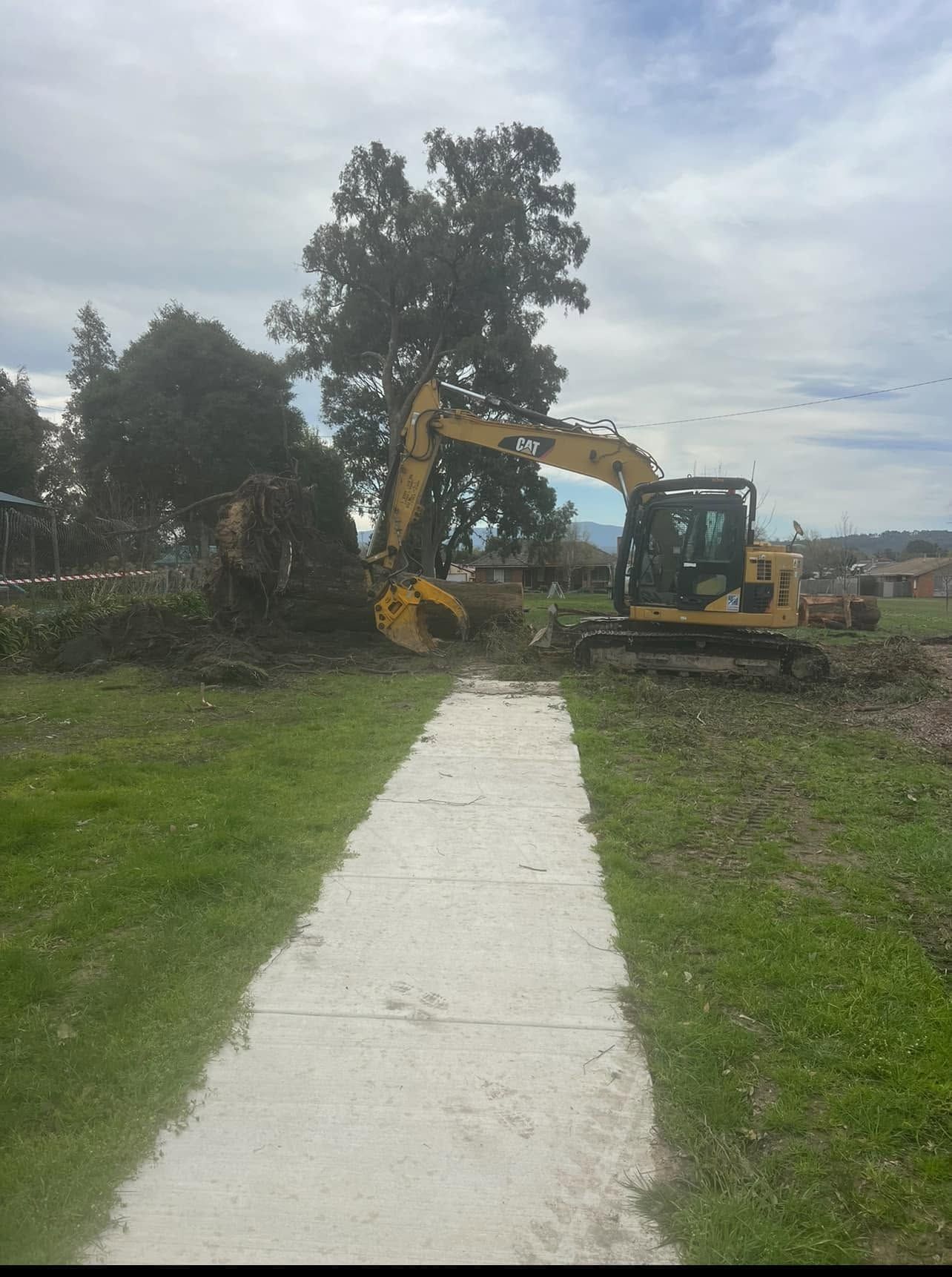 Yellow excavator clearing brush beside a sidewalk in a grassy area under cloudy skies