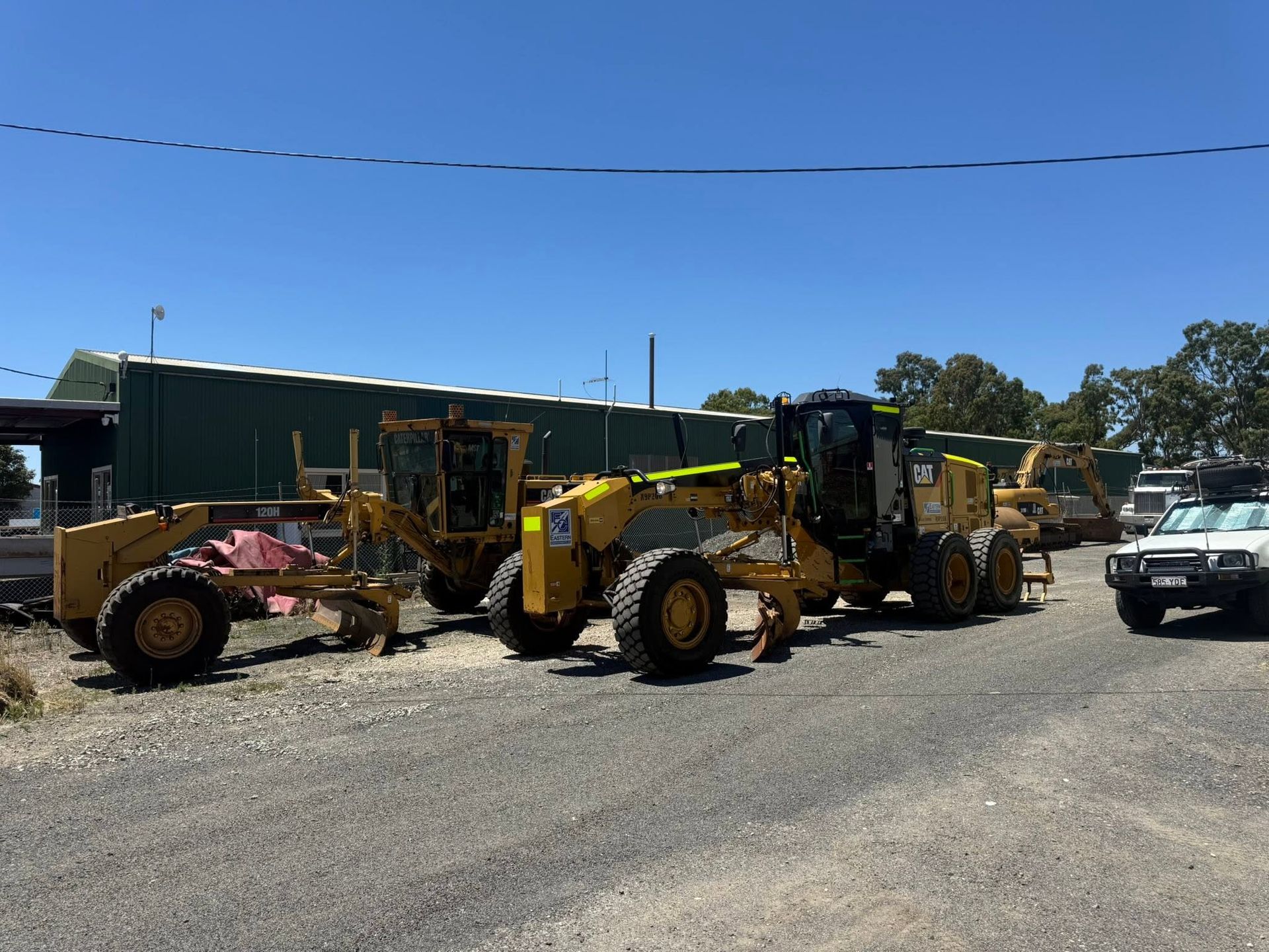 Yellow construction vehicles parked on gravel lot beside a green building under a blue sky