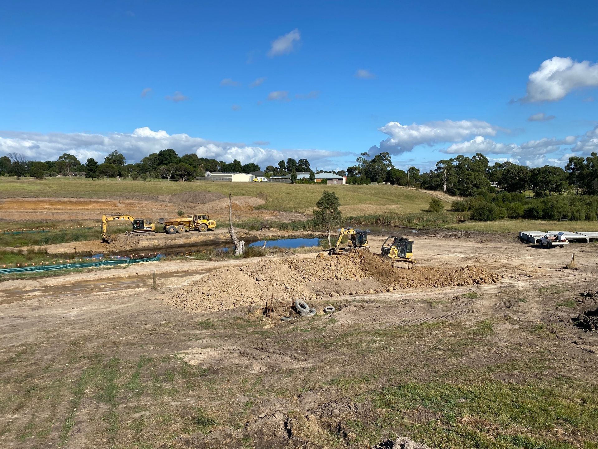 Yellow excavator on a grassy slope beside a house, digging red soil near a retaining wall.