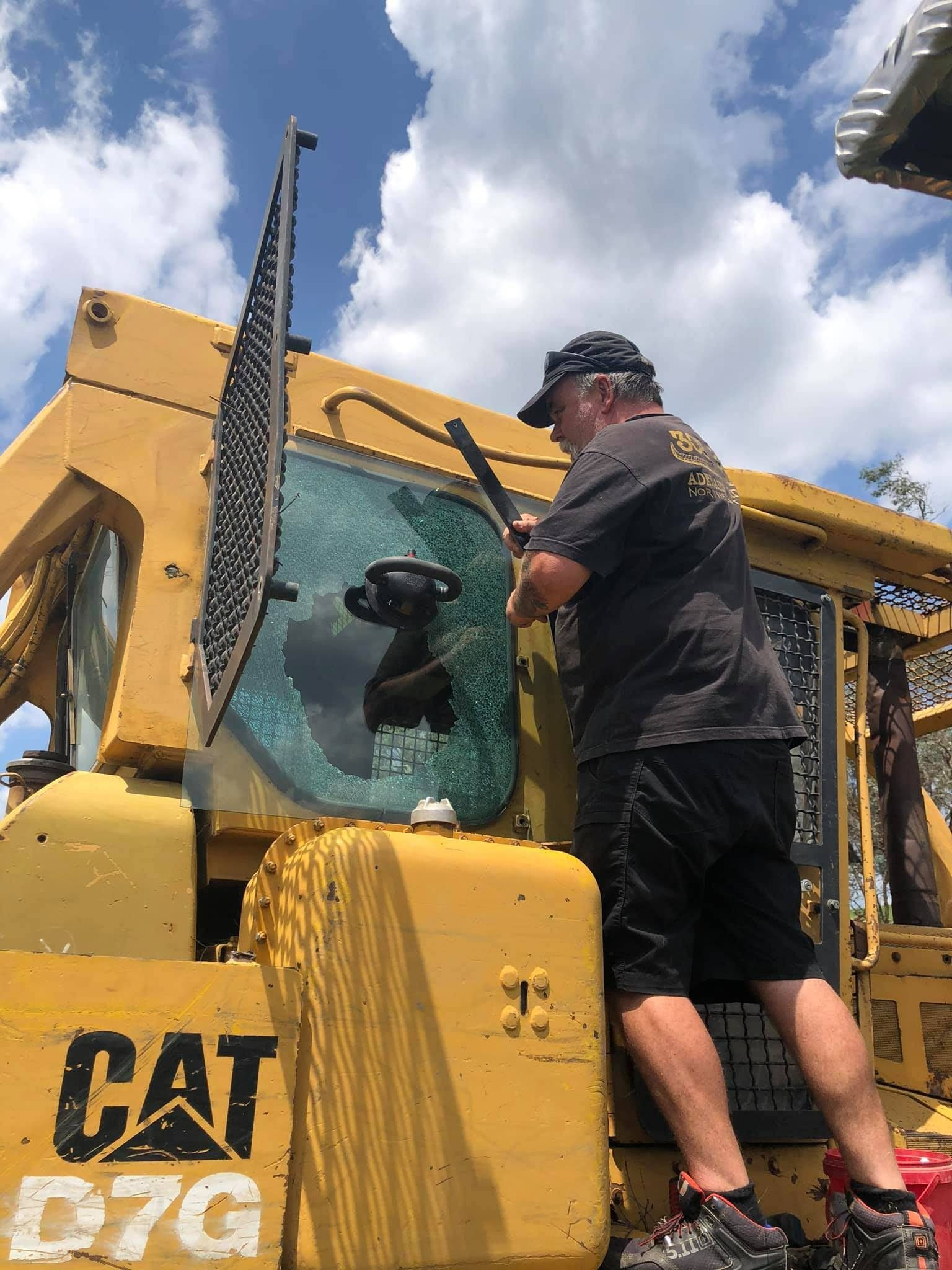 Man Smashes a Broken Window on a Yellow Cat D7g Bulldozer Under a Cloudy Sky — Road Runner Windscreens in South Grafton, NSW