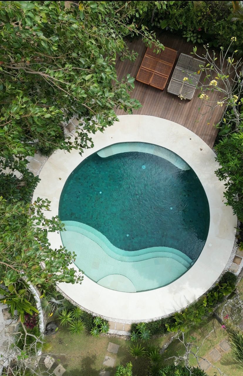 Overhead view of a round pool with turquoise water, surrounded by a white concrete deck and lush greenery.