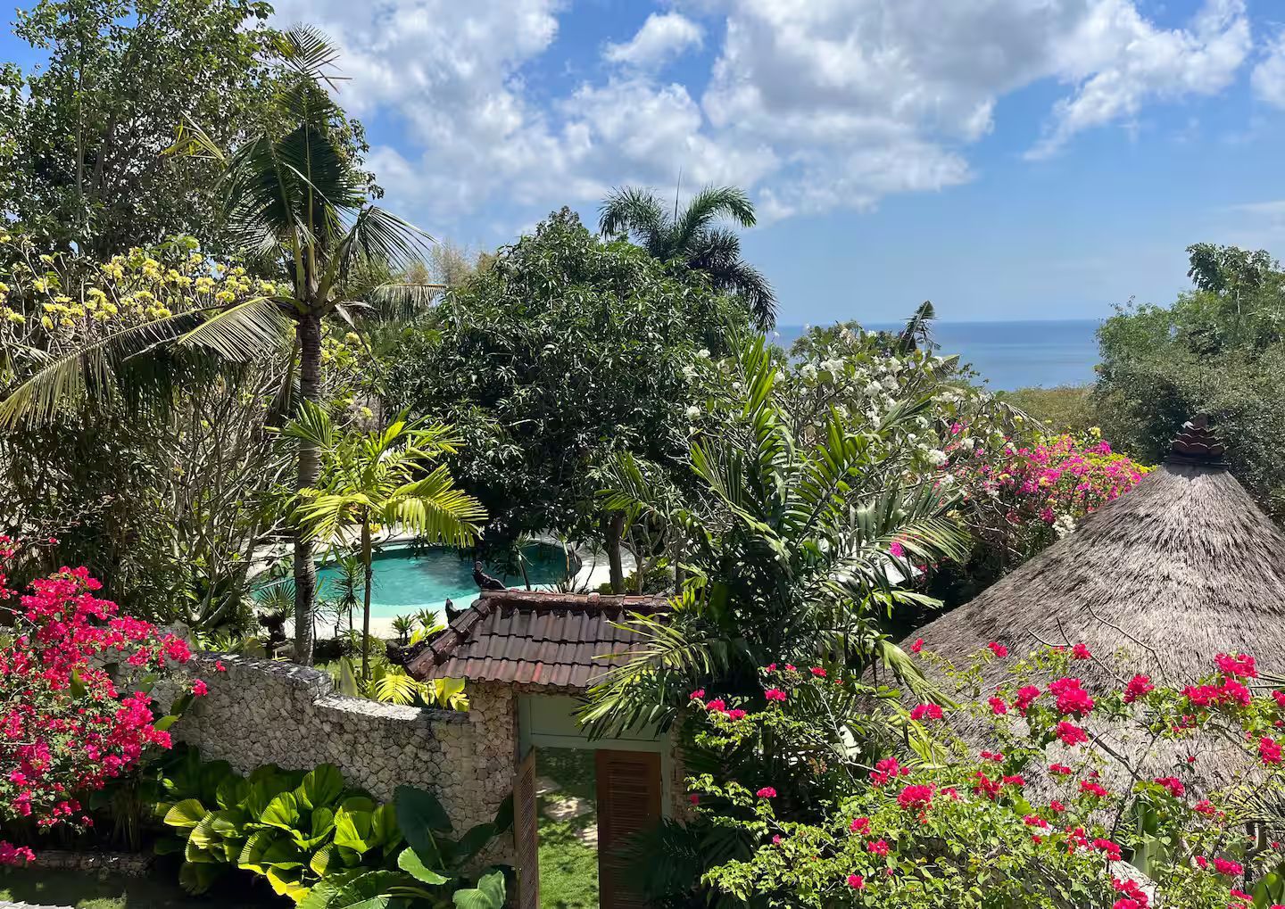 Overlooking a lush tropical garden with a pool, ocean view, and colorful blooms under a partly cloudy sky.
