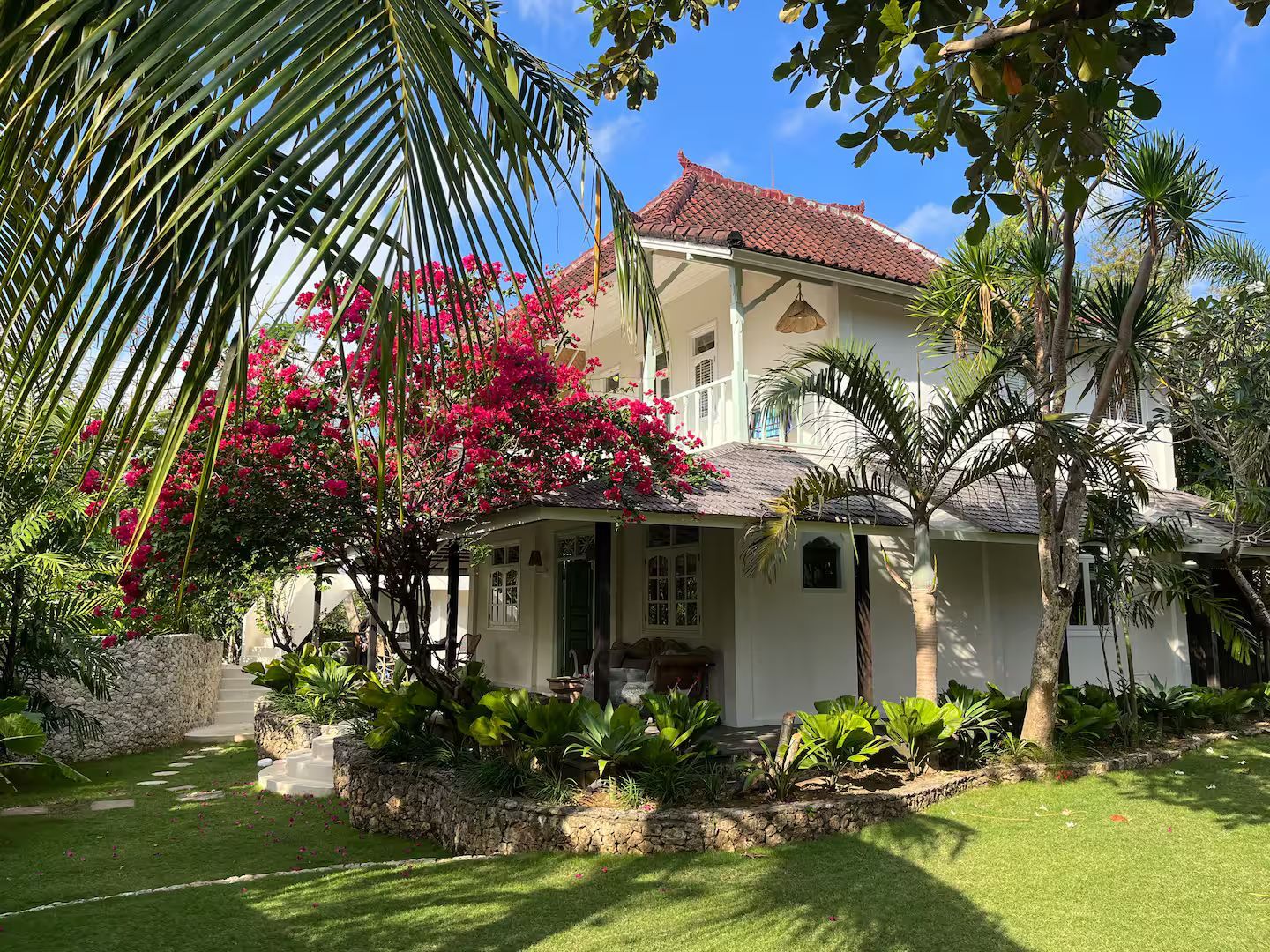 White two-story house with a red-tiled roof surrounded by lush greenery and a flowering bush in a sunny setting.