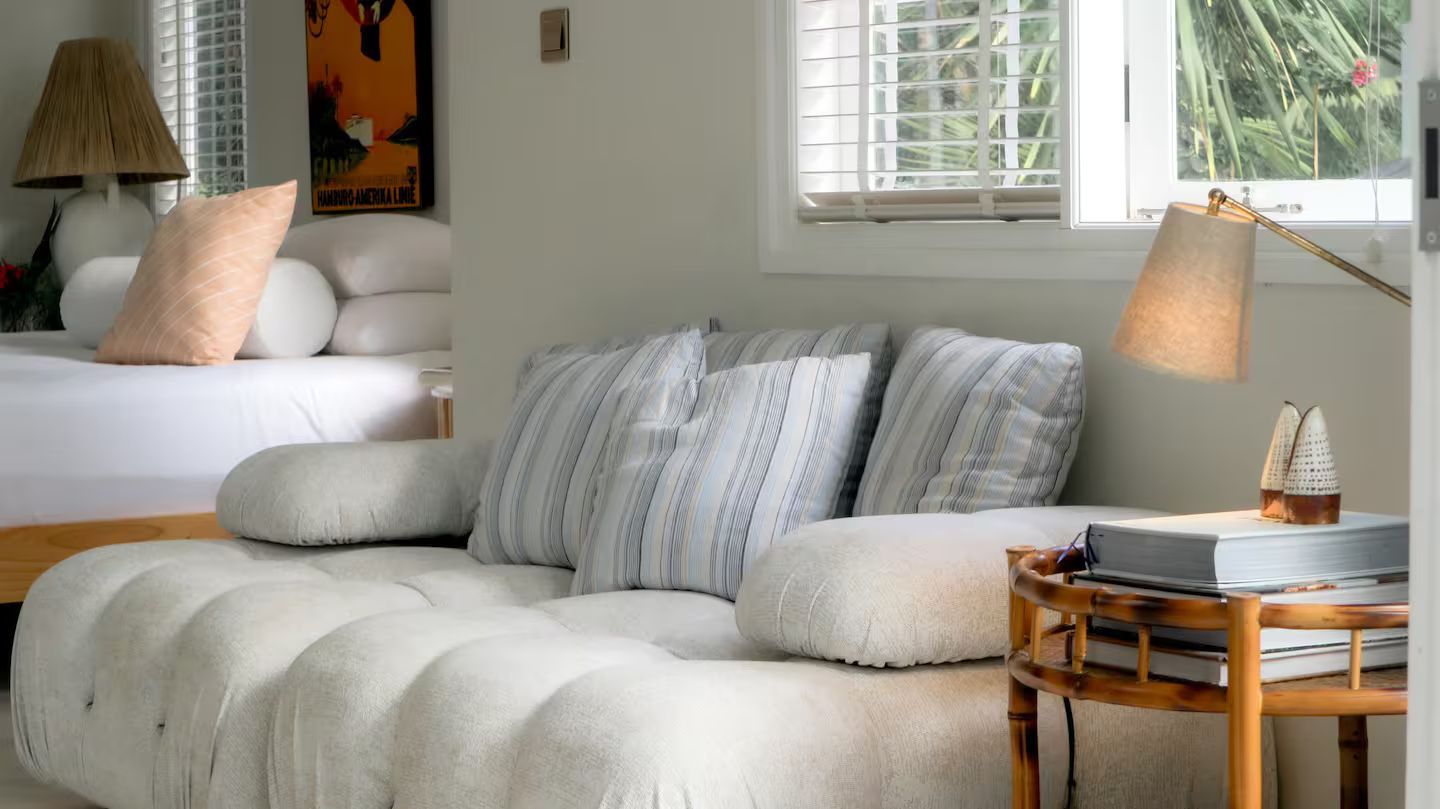 Cozy living space with a tufted sofa, striped pillows, and a bamboo side table with a lamp, near a window.