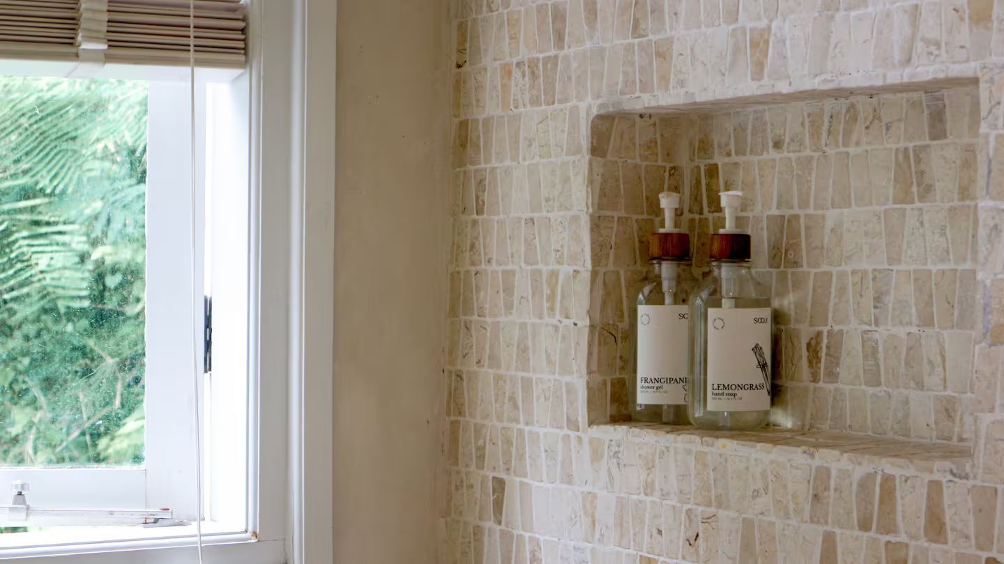A tiled shower niche with two shampoo bottles; a window with a bamboo blind is visible to the left.