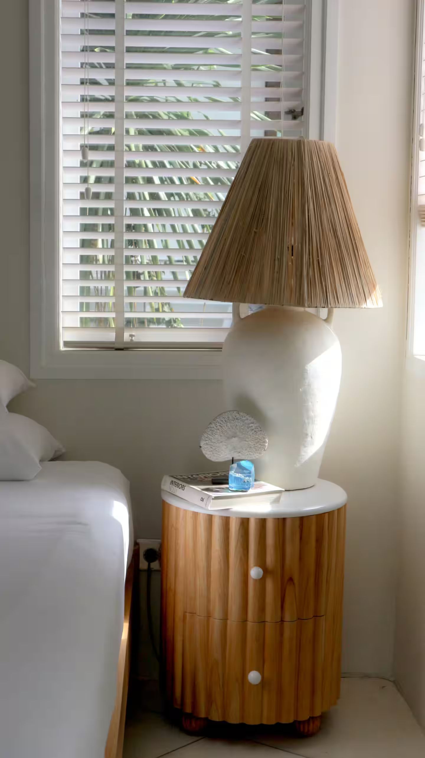 A bedside table with a large lamp, next to a bed and a window with closed blinds.