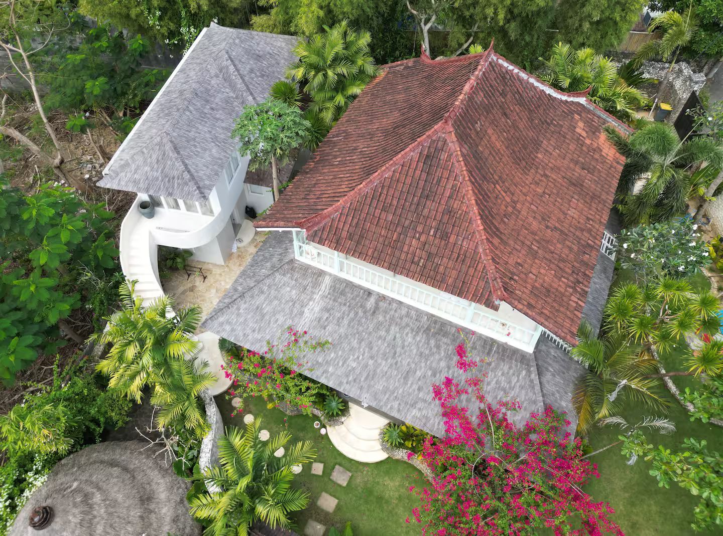 Aerial view of a white house with a terracotta-tiled roof and gray-tiled roof, surrounded by lush greenery and flowers.