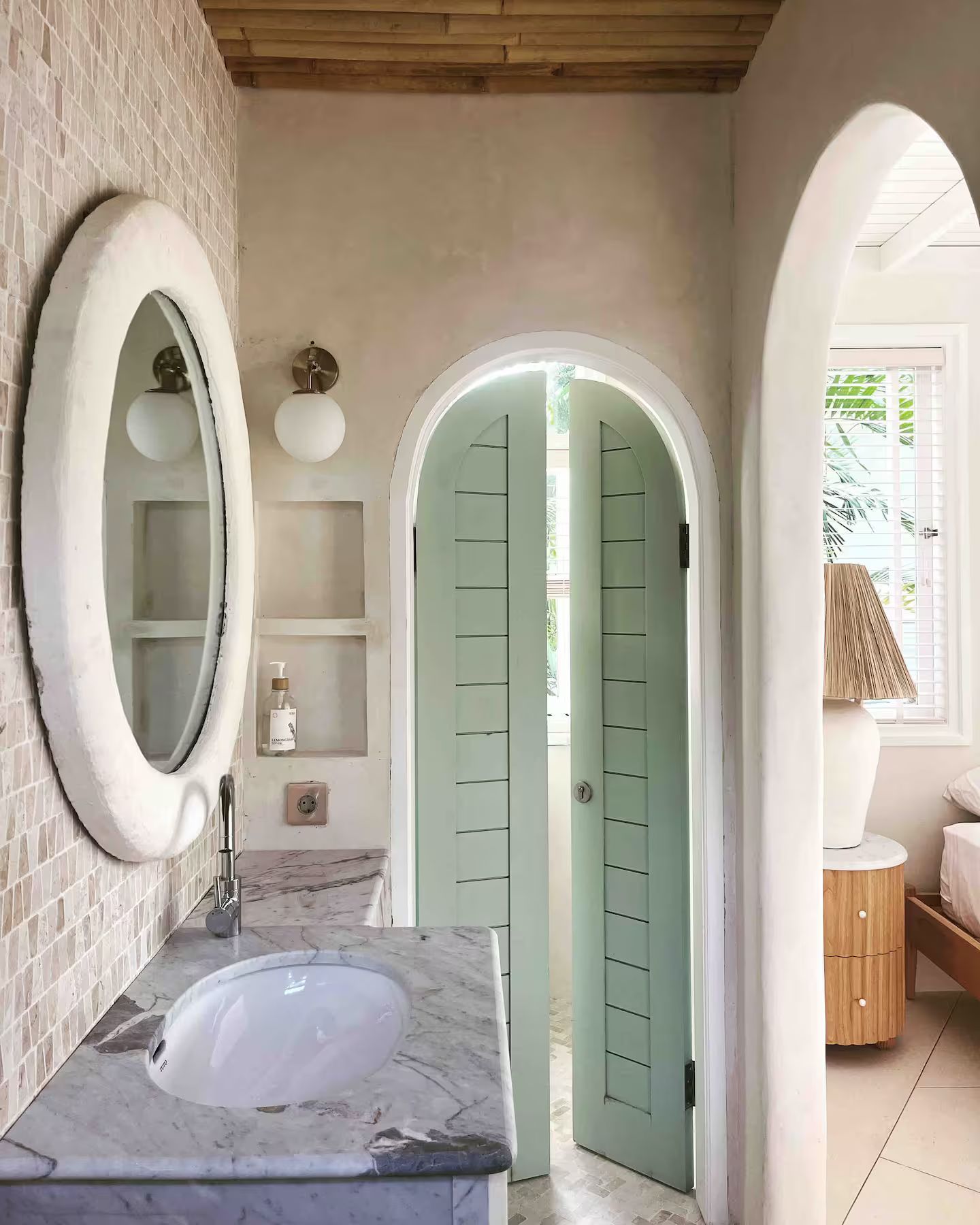 Bathroom with oval mirror over a marble vanity, leading to light green arched doors. Off-white walls and light wood accents.